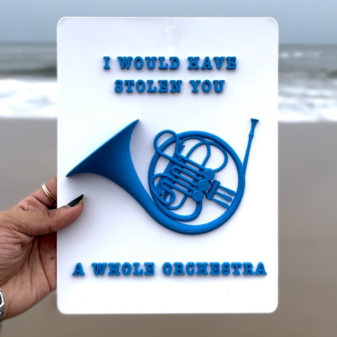 Card with blue French horn design and text held by a hand on a beach.